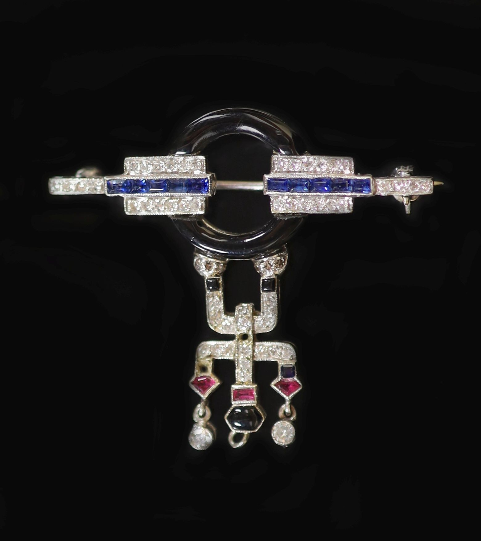 An Art Deco white gold, black enamel, ruby, sapphire and diamond set drop brooch